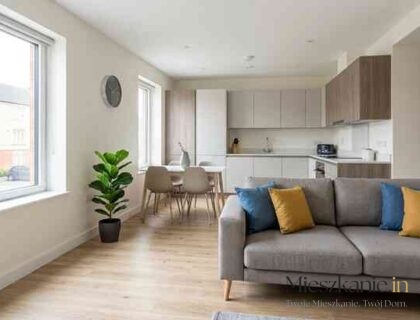 Nowoczesny, jasny salon z aneksem kuchennym wykończony w standardzie pod wynajem, z szarą kanapą i drewnianą podłogą.